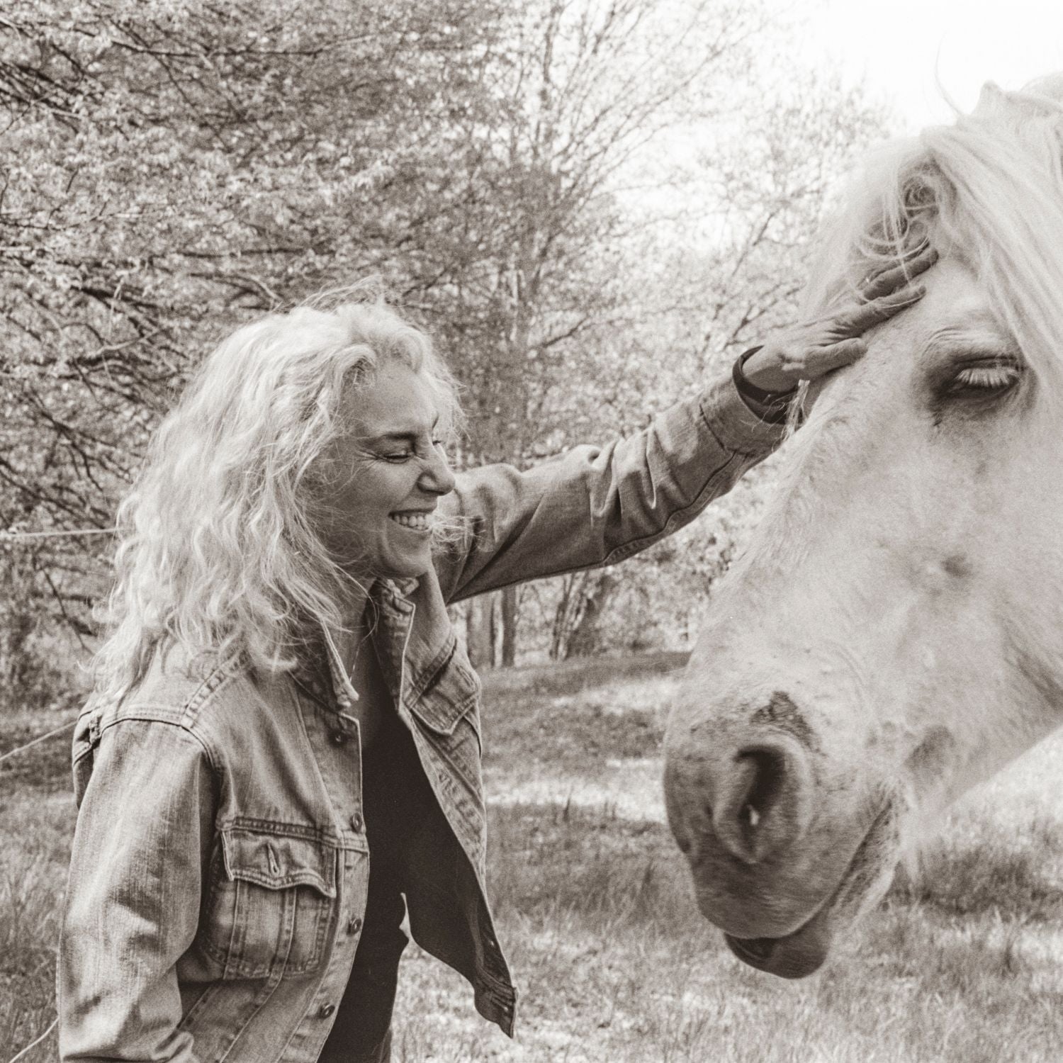 A woman with long curly hair laughs with her eyes closed as she rests her hand on the forehead of a white horse, in sepia.