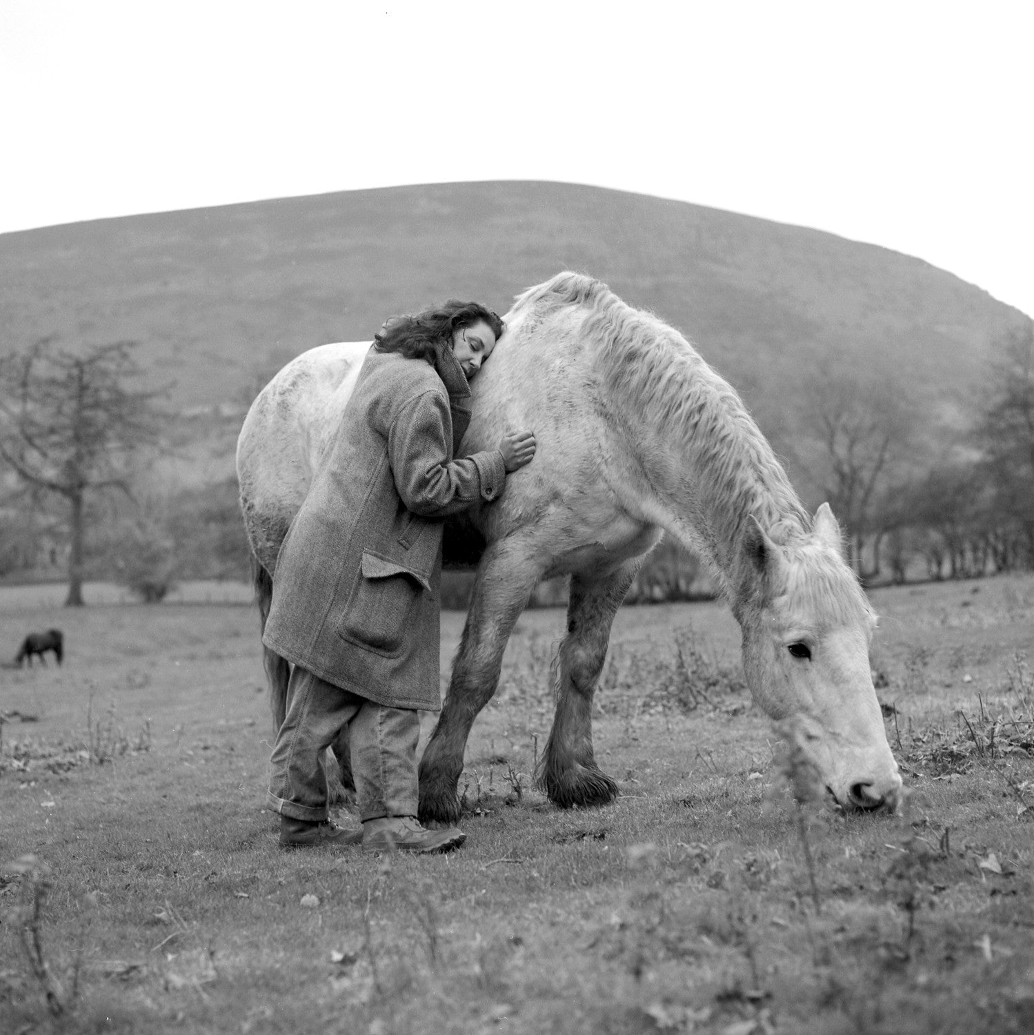 A woman in a tweed coat rests her head against the shoulder of a grazing white horse, with a bare hill behind them, in black and white.