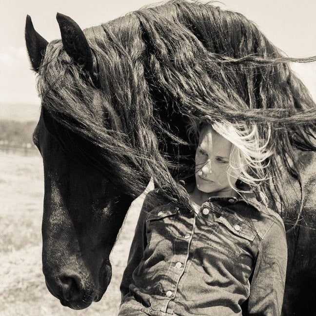 A woman rests her face against a dark horse's neck, its mane blown across her, in black and white.